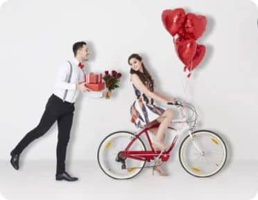 Homme offrant des fleurs à une femme en bicyclette, avec des ballons en forme de coeurs rouges accrochés au guidon